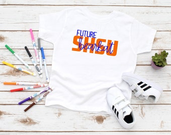 Könnte beinhalten: Weißes T-Shirt mit dem Text "FUTURE SHSU bearakt" in orangefarbener und blauer Schrift. Das Shirt liegt auf einer weißen Holzoberfläche mit Markern und einem Paar weißen Sneakers mit schwarzen Streifen.