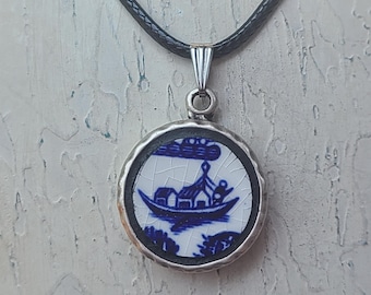 May include: A round pendant necklace featuring a blue and white porcelain design. The pendant depicts a boat scene with a silver-colored metal frame and bail. The necklace has a black cord.