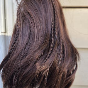 Op de afbeelding: Donkerbruine pruik met talrijke kleine vlechten door het haar. De pruik heeft een natuurlijke uitstraling met verschillende bruintinten. De vlechten zijn gelijkmatig verdeeld en voegen textuur toe. Het haar lijkt lang en vloeiend.