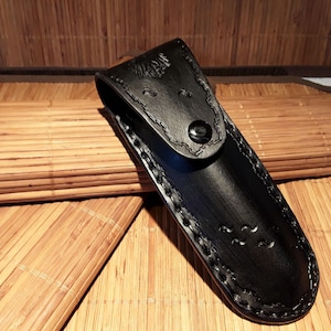 May include: A black leather sheath with decorative stitching and a snap closure. The sheath is designed to hold a knife or similar tool. The background is a bamboo mat.