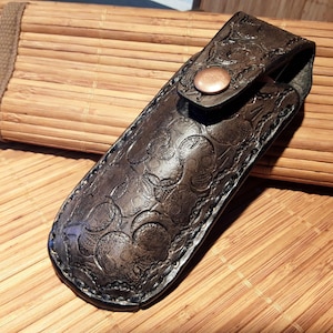 May include: A dark brown leather knife sheath with a decorative embossed pattern and a copper-colored snap closure. The sheath is designed to hold a knife and is shown on a bamboo mat.