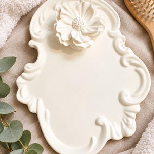 May include: An off-white ceramic decorative tray featuring an ornate, scrolled border and a three-dimensional sculpted flower centered at the top. The tray has a smooth, matte finish and a distinctively shaped, irregular silhouette with curved edges.