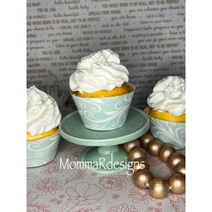 Op de afbeelding: Drie gele cupcakes met witte frosting worden gepresenteerd op een lichtgroene taartstandaard. De cupcakes zijn versierd met een lichtblauw en wit swirl-patroon. Een gouden kralenkrans ligt op de tafel onder de standaard.