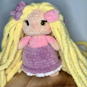 May include: A handmade plush doll with long yellow hair, a pink and purple dress, and flower accents. The doll has a light skin tone, black eyes, and green irises. The doll is made of soft, textured yarn.