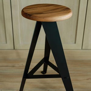May include: A modern bar stool with a round, wooden seat and black metal legs. The seat is made of a light brown wood. The legs are a matte black and form a triangular base for stability. The stool is designed for indoor use.