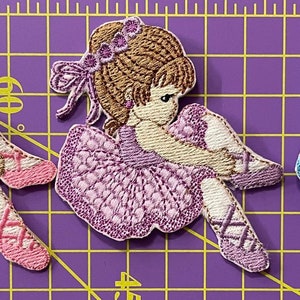 Può includere: Tre toppe ricamate di una ballerina in rosa, viola e blu. Ogni toppa presenta una ballerina in posa seduta, con un tutù e scarpette da ballo. Le toppe sono su uno sfondo a griglia viola.
