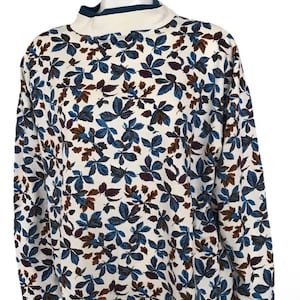 May include: A long-sleeved top featuring a white backdrop adorned with a pattern of blue, brown, and purple leaves. It has a high, white collar with a teal trim.