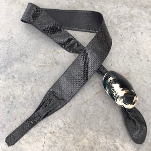 May include: A black snakeskin belt with a decorative shell buckle. The buckle is white and black with a textured surface.