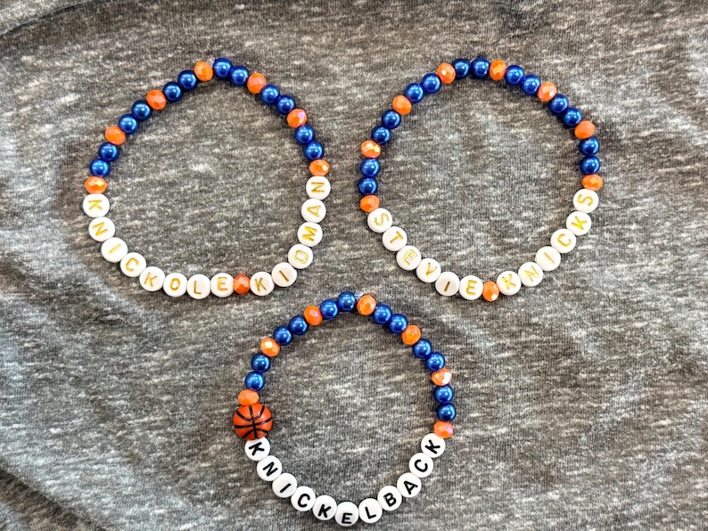 May include: Three beaded bracelets featuring blue and orange spherical beads. Two bracelets contain white letter beads spelling KNICKOLE KIDMAN and STEVIE KNOCKS. The third bracelet features white letter beads spelling KNICKELBACK and a basketball bead.