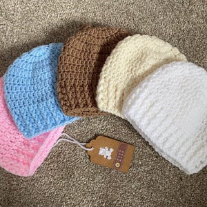 May include: A set of five crocheted beanies in pink, light blue, brown, cream, and white. Each beanie features a textured stitch pattern. A brown rectangular tag with a white bear graphic and the text HAND MADE is attached by a white string.