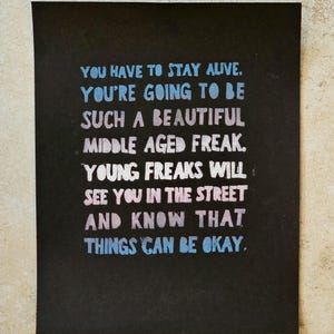 May include: A rectangular black print featuring the text: YOU HAVE TO STAY ALIVE. YOU'RE GOING TO BE SUCH A BEAUTIFUL MIDDLE AGED FREAK. YOUNG FREAKS WILL SEE YOU IN THE STREET AND KNOW THAT THINGS CAN BE OKAY. The lettering is in light blue, pink, and white.