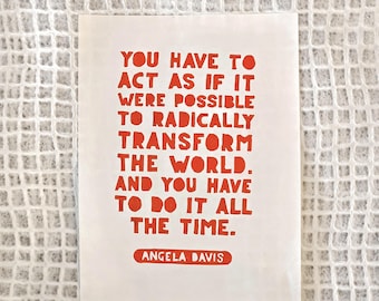 May include: A rectangular white print featuring bold, orange, hand-lettered text that reads: YOU HAVE TO ACT AS IF IT WERE POSSIBLE TO RADICALLY TRANSFORM THE WORLD. AND YOU HAVE TO DO IT ALL THE TIME. ANGELA DAVIS is printed below in an orange box.