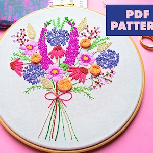 May include: A colorful embroidery hoop with a floral bouquet design. The bouquet features pink, purple, orange, and green flowers. The hoop is made of wood and has a white fabric background. The text "PDF PATTERN" is in blue and pink letters on a blue background.
