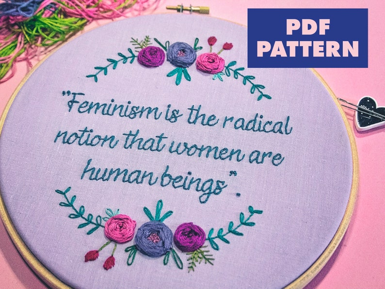 Puede incluir: Un aro de bordado morado con una cita que dice "El feminismo es la idea radical de que las mujeres son seres humanos." La cita est&aacute; rodeada de una corona de hojas verdes y flores rosas y moradas.