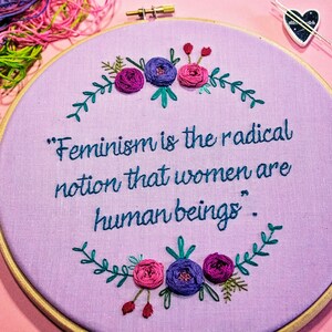 Puede incluir: Aro de bordado morado con una cita que dice "El feminismo es la idea radical de que las mujeres son seres humanos." La cita est&aacute; rodeada de un dise&ntilde;o floral con flores rosas y moradas.