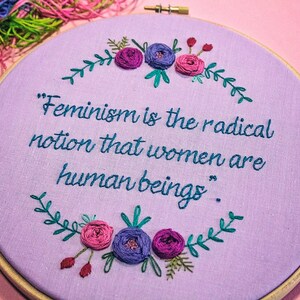 Puede incluir: Un aro de bordado morado con una cita que dice "El feminismo es la idea radical de que las mujeres son seres humanos." La cita est&aacute; rodeada de una corona de hojas verdes y flores rosas y moradas.