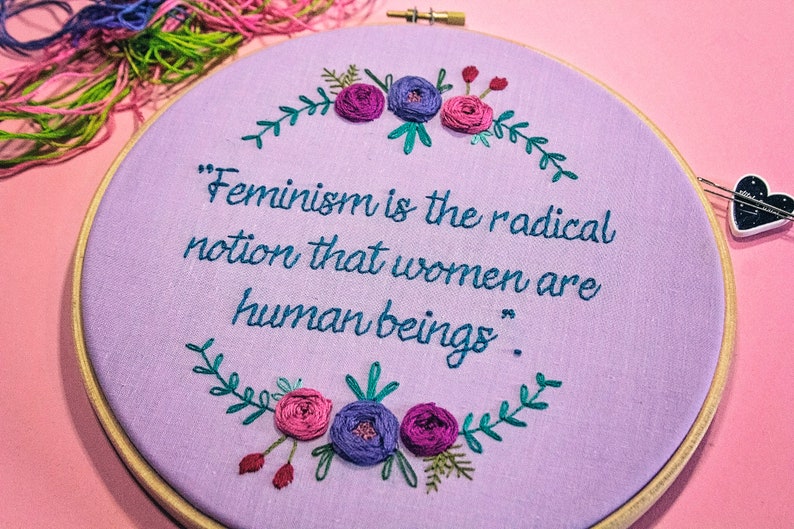 Puede incluir: Un aro de bordado morado con una cita que dice "El feminismo es la idea radical de que las mujeres son seres humanos." La cita est&aacute; rodeada de una corona de hojas verdes y flores rosas y moradas.