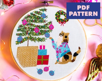 Könnte beinhalten: Gesticktes Hoop-Art mit einem Weihnachtsbaum, einer Katze im Pullover und Geschenken. Der Baum ist mit rosa und blauen Kugeln geschmückt. Die Katze trägt einen blauen Pullover mit Schneeflocken. Der Text "PDF PATTERN" ist zu sehen.
