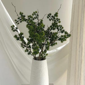 May include: A white textured vase holds a vibrant arrangement of green, needle-like branches. The vase has a slender, elongated shape. The background features soft, white fabric and a neutral-toned curtain, creating a minimalist aesthetic.