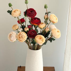 May include: A white textured vase holding a bouquet of artificial flowers. The flowers are cream, peach, and deep red ranunculus, with green stems and buds. The vase is on a wooden surface.