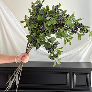 May include: A large artificial bouquet of dark blue berries on thin brown stems with numerous green leaves. The cluster of berries is dense and distributed across multiple branching stems, creating a full and rounded botanical arrangement.