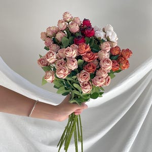 May include: A bouquet of artificial roses in various shades of dusty pink, deep red, burnt orange, and cream. The flowers feature layered petals and green leaves on thin stems. The arrangement is gathered together in a dense, rounded cluster.