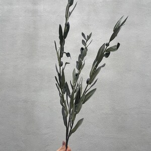 May include: A sprig of faux olive branches with dark green leaves and small, dark olive-like berries. The artificial plant is held against a light gray background, showcasing its naturalistic design for home decor.