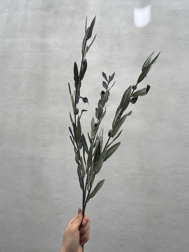 May include: A sprig of faux olive branches with dark green leaves and small, dark olive-like berries. The artificial plant is held against a light gray background, showcasing its naturalistic design for home decor.