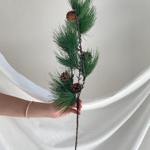 May include: A decorative artificial pine branch featuring long, slender green needles and four brown pinecones attached to a thin, dark brown stem. The branch is structured with multiple smaller sprigs extending from the main central stalk.