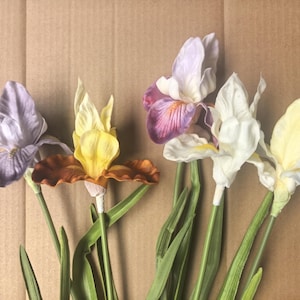 May include: A collection of four artificial iris flowers with green stems and leaves. The flowers feature varied color palettes including light purple, a combination of yellow and brown, a mix of purple and white, and a pale yellow and blue blend.
