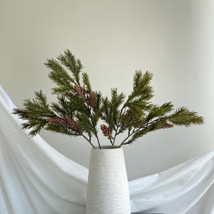 May include: A white ceramic vase with a textured, ridged surface holds several artificial pine branches. The branches feature deep green needles and small, light brown pinecones distributed throughout the foliage, creating a natural arrangement.