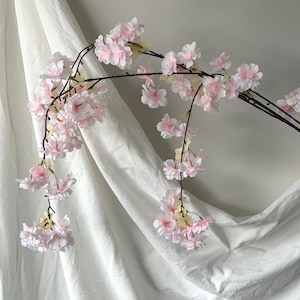 May include: A branch of artificial cherry blossoms with delicate pink petals and yellow centers. The blossoms are attached to dark brown stems, creating a naturalistic look. The flowers are set against a white backdrop.