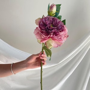 May include: A single artificial peony flower stem featuring a large, multi-layered bloom with deep purple and soft pink petals, a smaller unopened bud at the top, and several green leaves attached to a long, slender green stem.