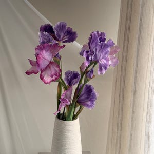 May include: A white textured vase holds a bouquet of purple and pink iris flowers. The irises have ruffled petals and green stems. The vase is on a white surface, with a sheer white curtain and a striped curtain in the background.