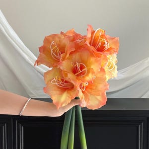 Puede incluir: Un ramo de flores de amarilis artificiales con grandes pétalos en forma de trompeta en tonos vibrantes de naranja y amarillo. Las flores tienen estambres blancos prominentes con puntas amarillas y están unidas a tres tallos verdes gruesos.