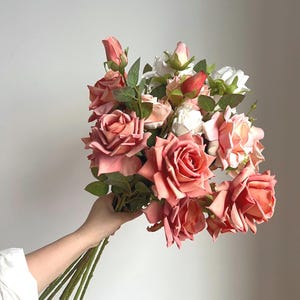Könnte beinhalten: Ein Strauß künstlicher Rosen in Rosa-, Pfirsich- und Weißtönen. Das Arrangement umfasst voll erblühte Rosen, Knospen und grüne Blätter. Die Stiele sind lang und grün.