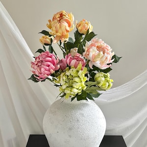 Op de afbeelding: Een boeket kunstmatige chrysanten in tinten perzik, roze en limoengroen, geschikt in een ronde, getextureerde witte vaas. De bloemen hebben gelaagde, gekrulde bloemblaadjes en donkergroene bladeren aan de stelen voor een levendig geheel.