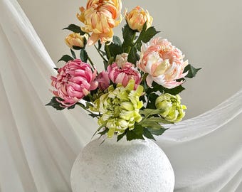 Può includere: Un bouquet di crisantemi artificiali nelle tonalità pesca, rosa e verde lime, disposto in un vaso rotondo bianco dalla superficie ruvida. I fiori presentano petali arricciati a strati e foglie verde scuro sui gambi.