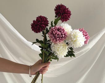 Può includere: Un bouquet di crisantemi artificiali con fiori color bordeaux intenso, rosa tenue e bianco crema. I fiori presentano petali densi e stratificati e sono fissati su lunghi steli verdi sottili con piccole foglie verdi lungo i gambi.