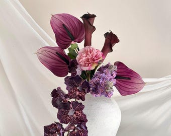 May include: A white textured ceramic vase containing a floral arrangement of deep purple anthuriums, dark calla lilies, a single light pink peony, a cluster of light purple lilacs, and a cascading stem of dark purple spotted orchids.