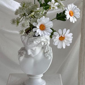 May include: A white ceramic bust vase holding white daisies and greenery. The vase is sculpted in the form of a classical head. The daisies have white petals and orange centres. The vase is on a white surface.