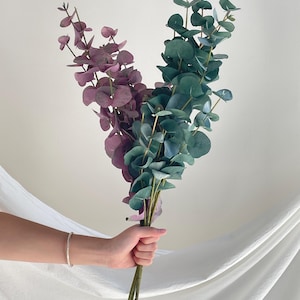Peut inclure: Un bouquet de tiges d'eucalyptus artificielles avec des feuilles arrondies dans des tons de violet sourd et de vert sarcelle profond. Les tiges sont longues et fines, se ramifiant pour soutenir le feuillage, créant un arrangement botanique vertical.