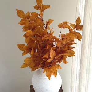May include: A white textured vase holds a bouquet of orange-brown leaves. The vase sits on a dark wooden surface. The leaves have a maple leaf shape and are clustered together, creating a fall-themed arrangement.
