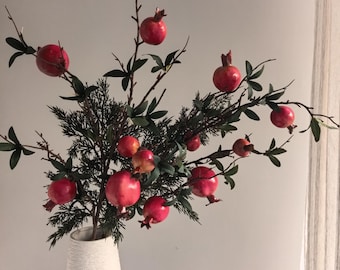 May include: A white textured vase holds a decorative arrangement of branches with vibrant red pomegranates and green leaves. The vase is set against a neutral background, creating a striking contrast with the colorful fruit.