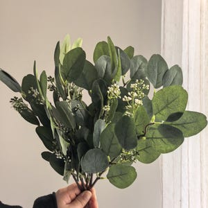 May include: A bundle of faux eucalyptus leaves with small white flower buds. The leaves are various shades of green, with rounded shapes and a slightly waxy appearance. The stems are brown and tied together, creating a natural, botanical aesthetic.
