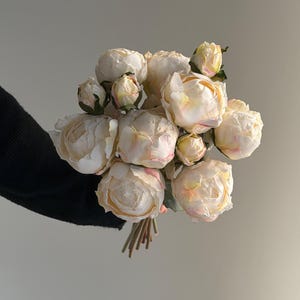 Puede incluir: Un ramo de peonías artificiales con múltiples flores abiertas y capullos cerrados en tonos crema y rosa pálido. Las flores tienen pétalos redondeados y superpuestos con bordes sutilmente teñidos de rosa, unidos por los tallos en la base.
