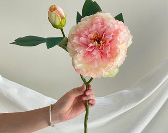 May include: A single artificial flower stem featuring one large, multi-layered peony bloom in shades of soft pink and cream, accompanied by a smaller, closed bud and several dark green leaves on a slender, textured green stalk.