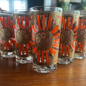 May include: Set of six vintage highball glasses. Each glass features a gold and orange design with floral and geometric patterns. The glasses are clear and have a classic, elegant look.