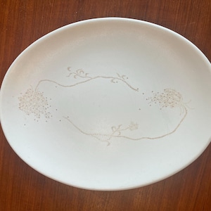 May include: A white oval-shaped ceramic plate with a delicate floral design. The design features a series of dotted clusters and connecting lines in a light beige color, creating a subtle, elegant pattern. The plate is set against a dark wooden surface.