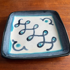 May include: Square ceramic plate with a white center and a teal and navy blue border. The plate features a repeating abstract design in navy blue. The plate is on a wooden surface.